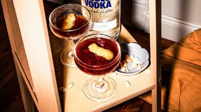 Black Martini in two coupe glasses with lemon peel garnishes on a wooden tray beside a vodka bottle and bowl of peanuts