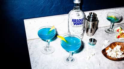 Two Blue Cosmopolitan cocktails garnished with lemon zest, next to a bottle of Absolut Vodka and a boston shaker.