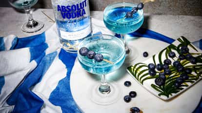 Blueberry Martini in coupe glasses with Absolut Vodka bottle and blueberry skewers on a marble board