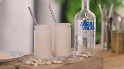 Cashew Hardshake in two glass jars with straws on wood with ice and cashews beside an Absolut Vodka bottle