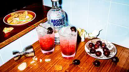 Dirty Shirley in two highball glasses with ice and cherries beside an Absolut Vodka bottle on a wood counter