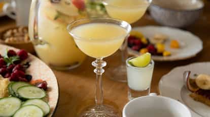 A pale yellow cocktail in a stem coupe with a small lime-garnished shot on a wooden table.