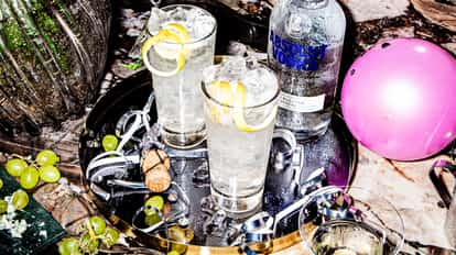 Two French 76 cocktails served in Highball Glasses filled with ice and garnished with a lemon twist on a metal tray surrounded by party streamers, balloons and a bottle of Absolut Vodka.