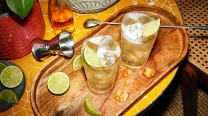 Gin Buck in two highball glasses on a wooden tray with ice, lime wedges and a metal jigger