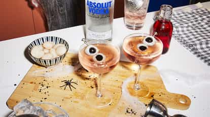 Lychee Eyeball Martini in coupe glasses with skewered lychees on a wooden board with an Absolut bottle behind