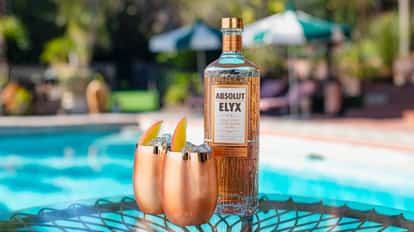 Elyx ManGo two metal tumblers with ice and mango wedges beside a vodka bottle on a glass table by a pool