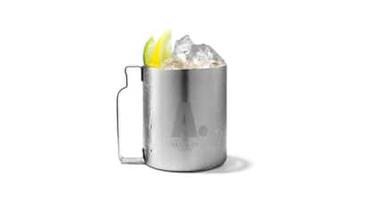 A cocktail in an Absolut metal mug is garnished with lemon and lime wedges on white background.