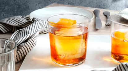 Two beautiful Old Fashioned cocktails served in a Rock Glass on a marble chopping board, one cocktail is slightly out of the frame, both with a large Ice Cube and with fresh Orange Zest.
