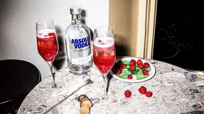 Raspberry Bellini served in two champagne flutes with raspberries and an Absolut Vodka bottle on a marble table