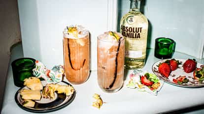 Two long glaases of strawberry night cocktail on a table with Absolut Vanilia bottle and  plates with sliced pine apple and strawberries.