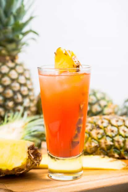 Zombie in a highball glass with pineapple wedge garnish on a wooden board with pineapple halves in background